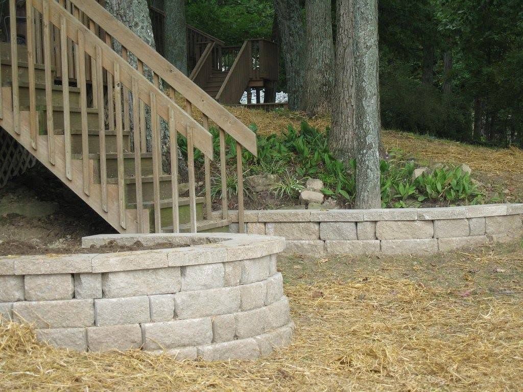Curved stone block planter and low retaining wall installed next to a wooden outdoor staircase on a wooded slope