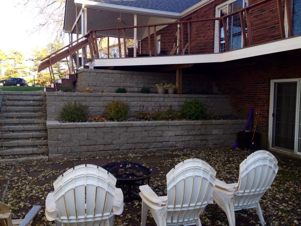 White Adirondack chairs around a fire pit below a multi-tiered gray block retaining wall and elevated wood deck