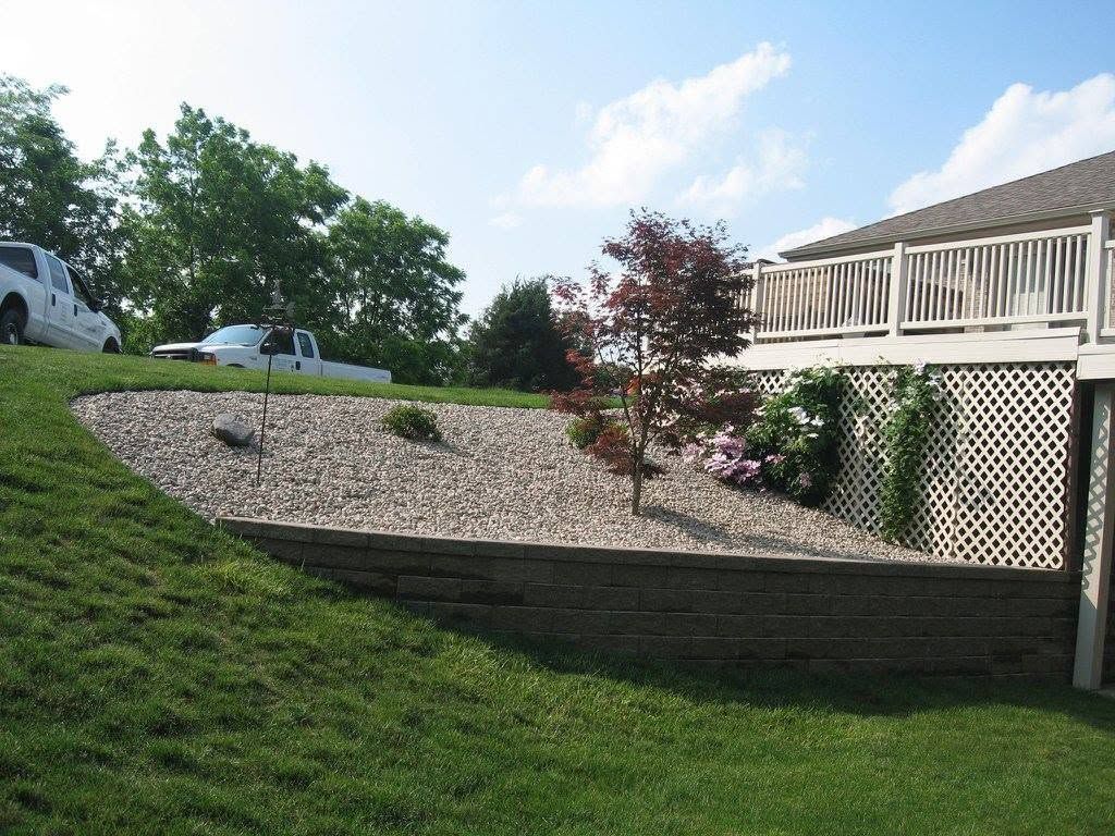 Sloped backyard landscaping featuring a dark block retaining wall, light gravel bed, and a small ornamental red maple tree