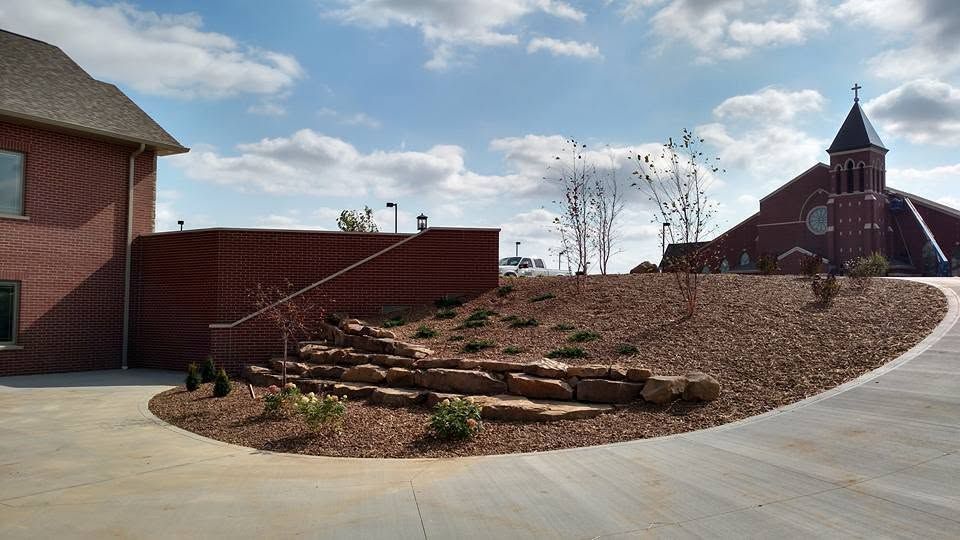 Multi-tiered natural stone retaining wall on a mulched hill with a brick structure and church steeple in the background