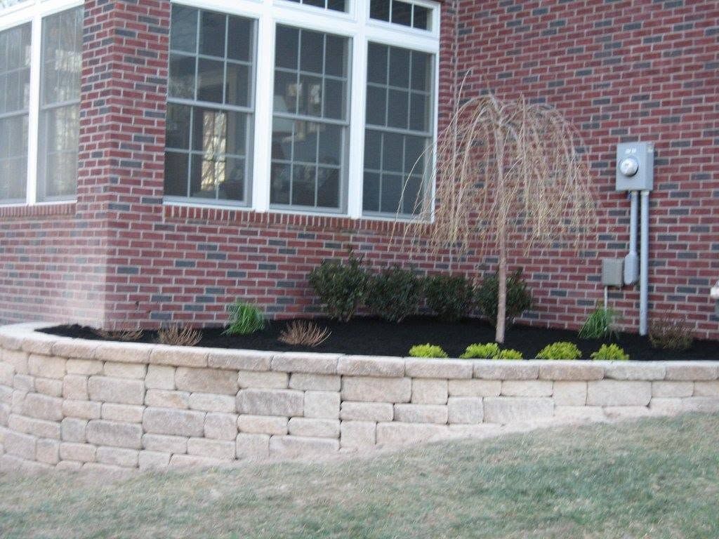 Light-colored stacked stone retaining wall with dark mulch and a small weeping tree in front of a red brick house