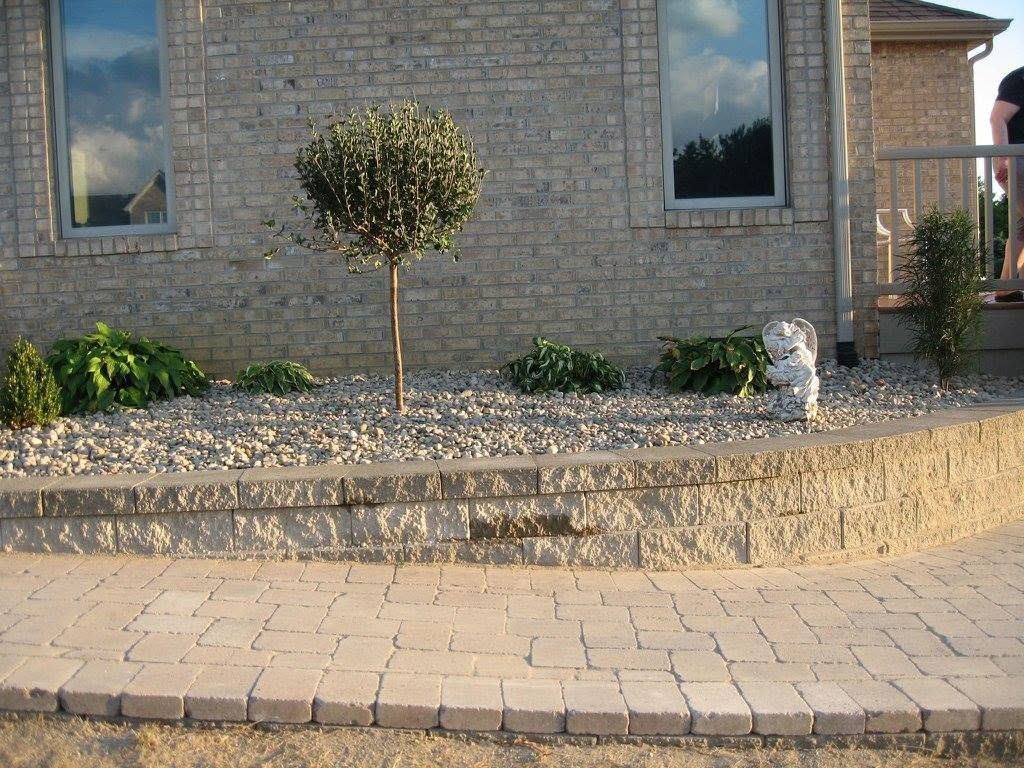 Brick paver walkway next to a raised stone block retaining wall with a small topiary tree and gravel ground cover