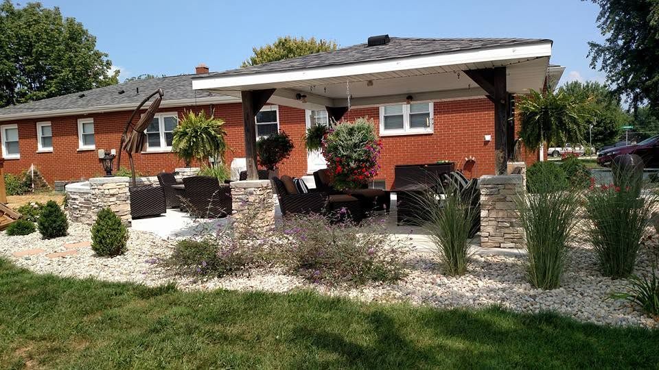 Covered backyard patio with stone-wrapped pillars, outdoor furniture, and hanging flower baskets attached to a red brick home