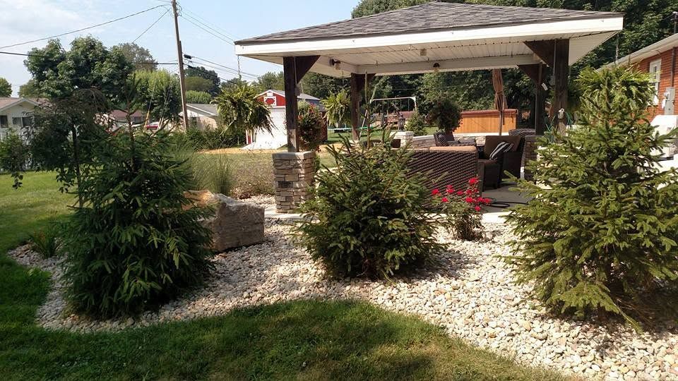 Covered outdoor living pavilion with stone-wrapped pillars and a landscaped gravel bed featuring an evergreen shrub