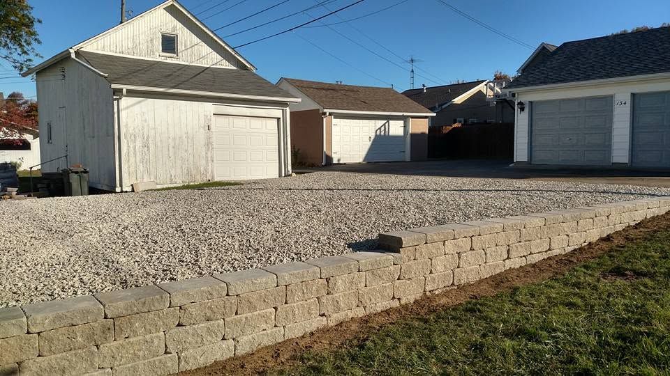 Long, low gray stacked block retaining wall bordering a gravel parking area beside detached garages