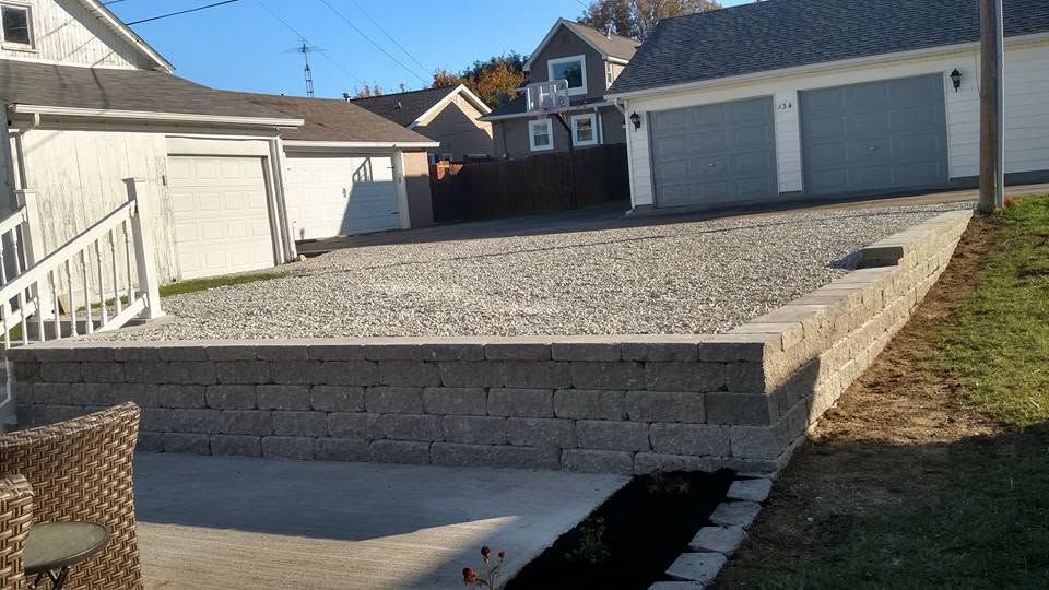 Gray stone block retaining wall separating a large gravel driveway from a lower concrete pad near residential garages