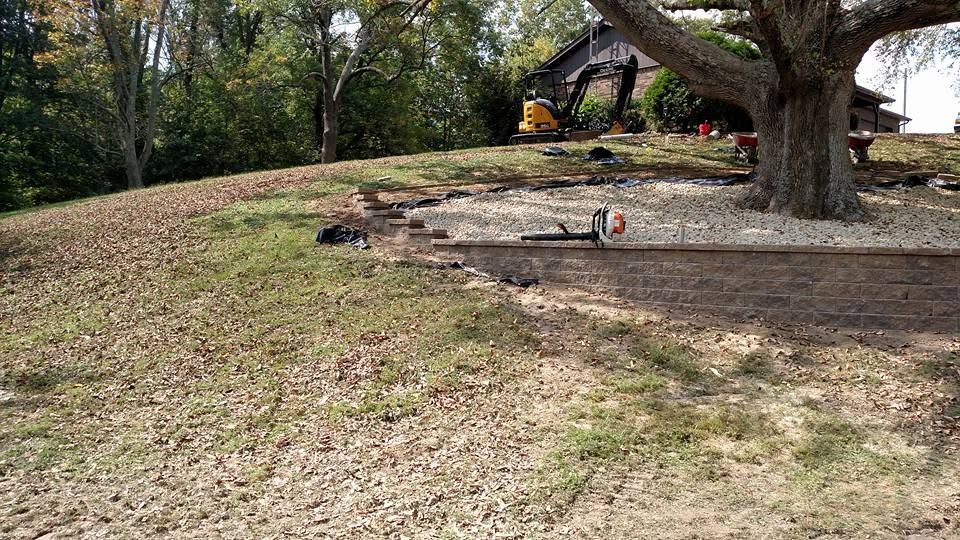 Stone block retaining wall under construction on a grassy hill with a mini excavator working in the background