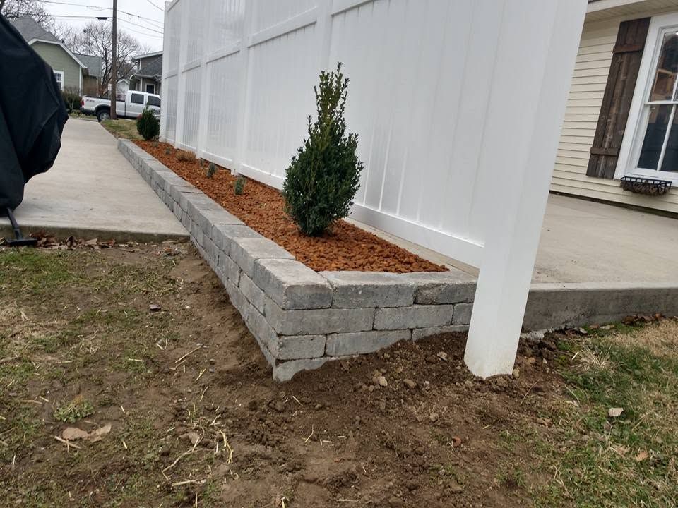 Long, raised gray stone planter bed with fresh mulch and an evergreen shrub alongside a white privacy fence