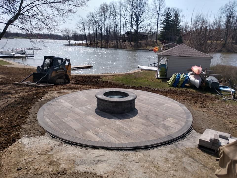 Circular paver patio construction with a stone fire pit overlooking a lake property.