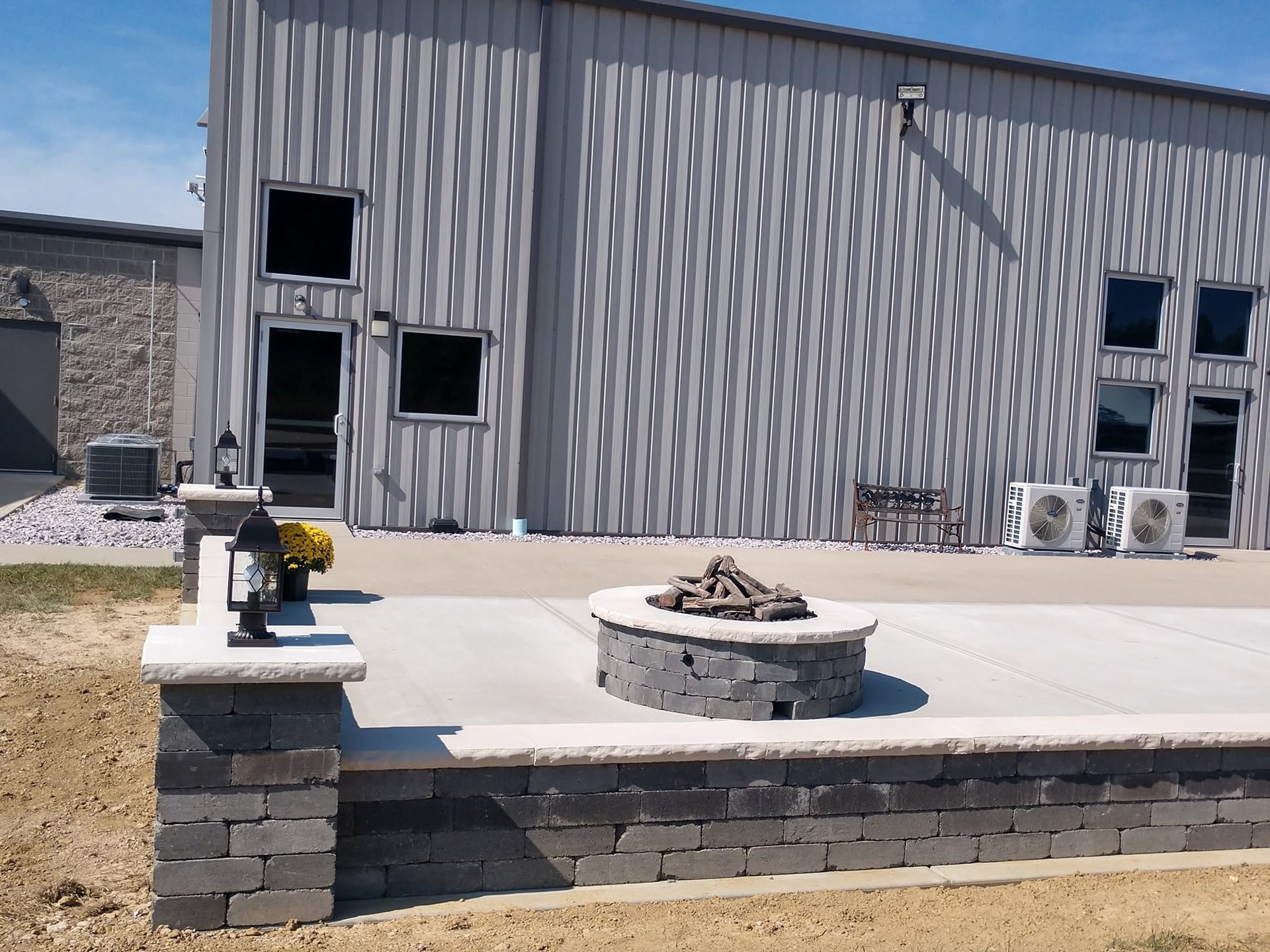 Circular stone block fire pit on a concrete pad enclosed by a low stone retaining wall outside a corrugated steel building