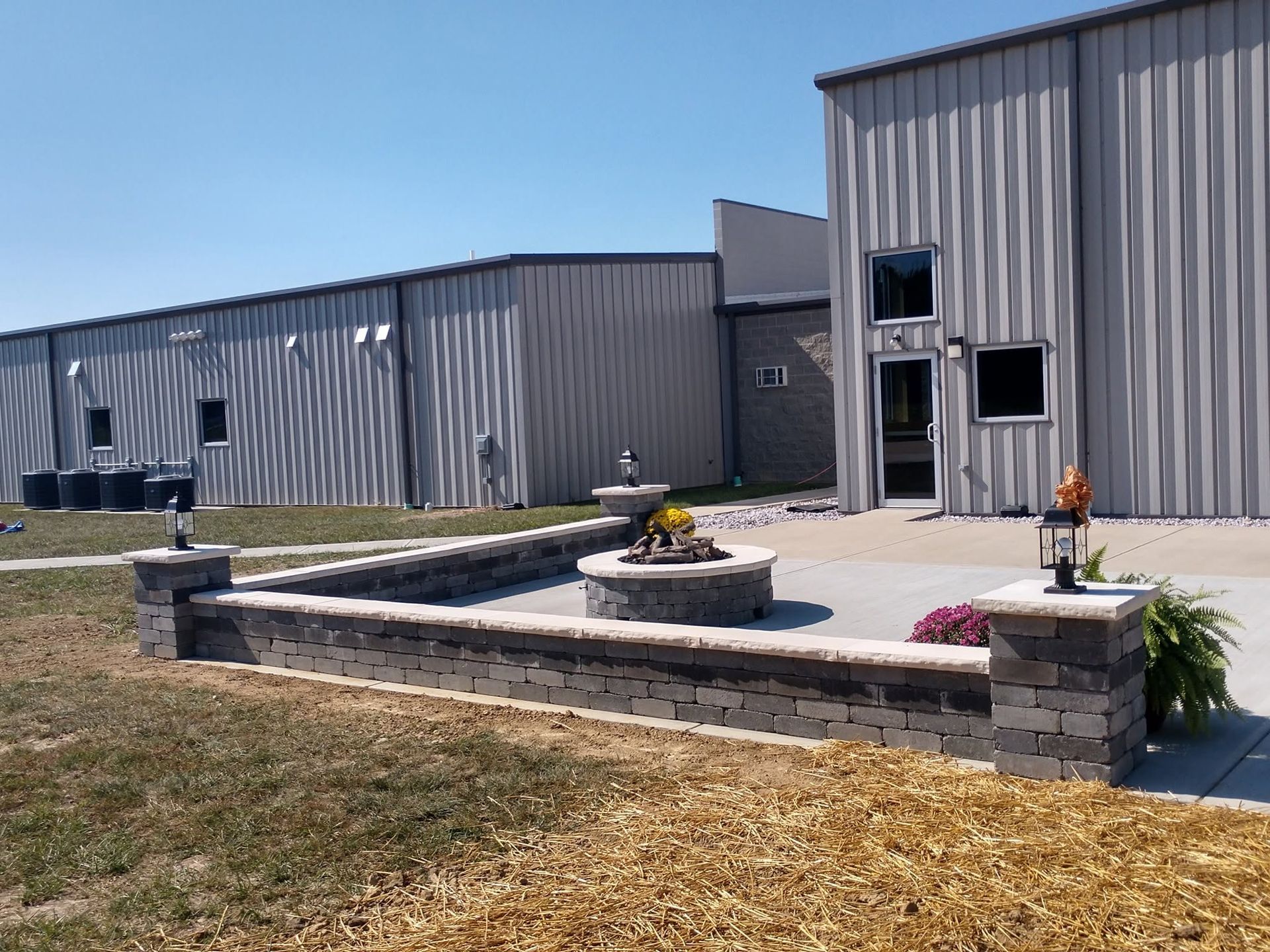 Spacious concrete patio with a circular stone fire pit and low seating wall installed next to a commercial metal building
