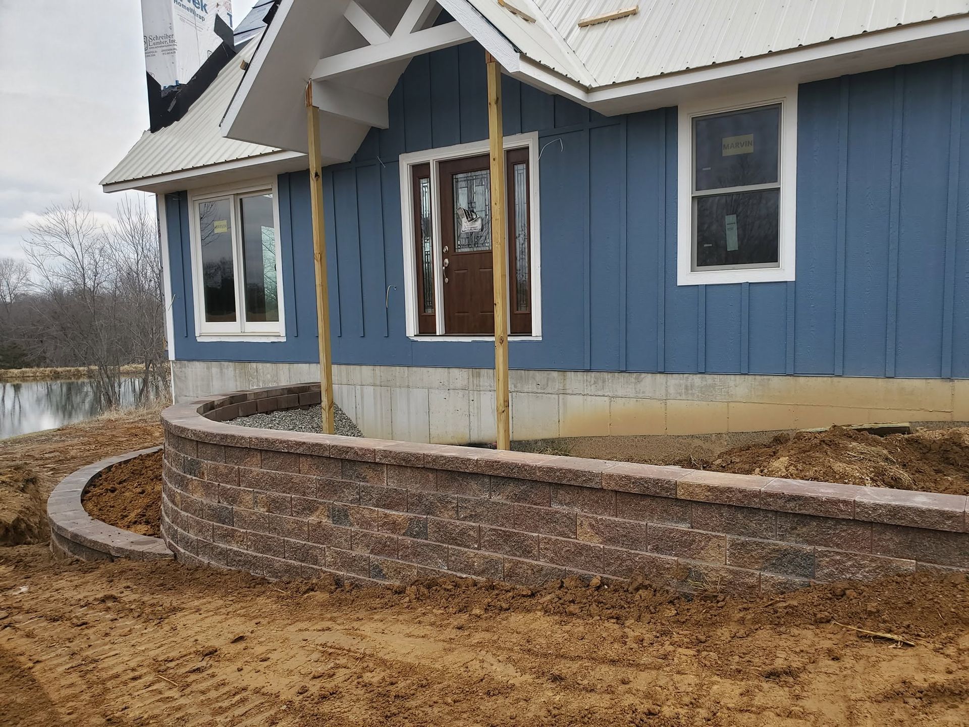 Tiered stone block retaining wall under construction along the foundation of a modern blue home.