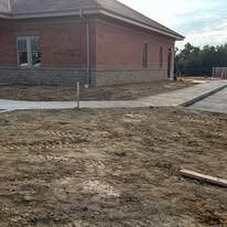 Site preparation and dirt grading work in progress along the side of a brick building and concrete walkway