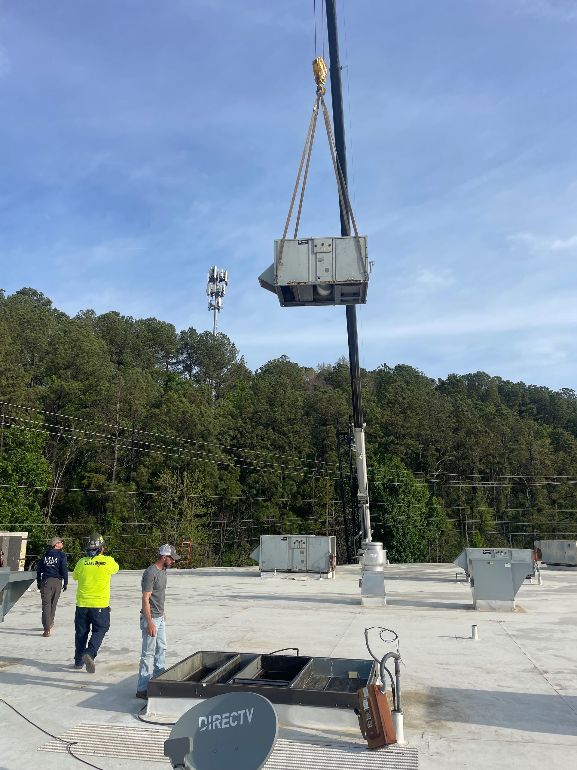 Crane lifting equipment onto a rooftop; workers observe. Antenna tower in background, sunny day.