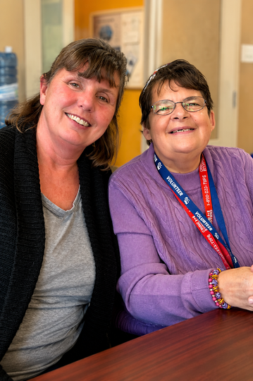 Two people smiling at a table indoors, one in a purple sweater with a lanyard.