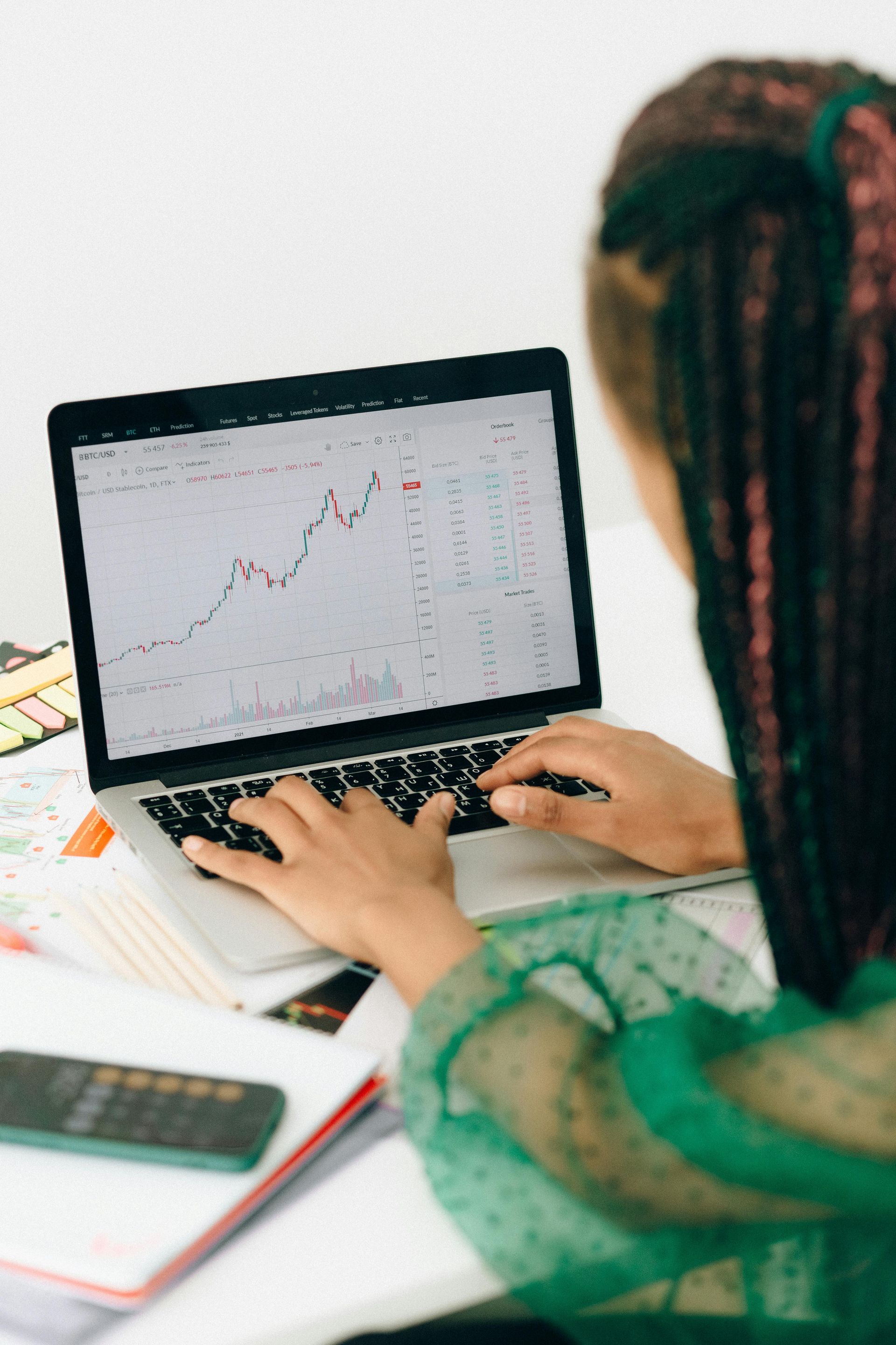 Woman with braided hair using a laptop, viewing a stock chart. Green top, white background.