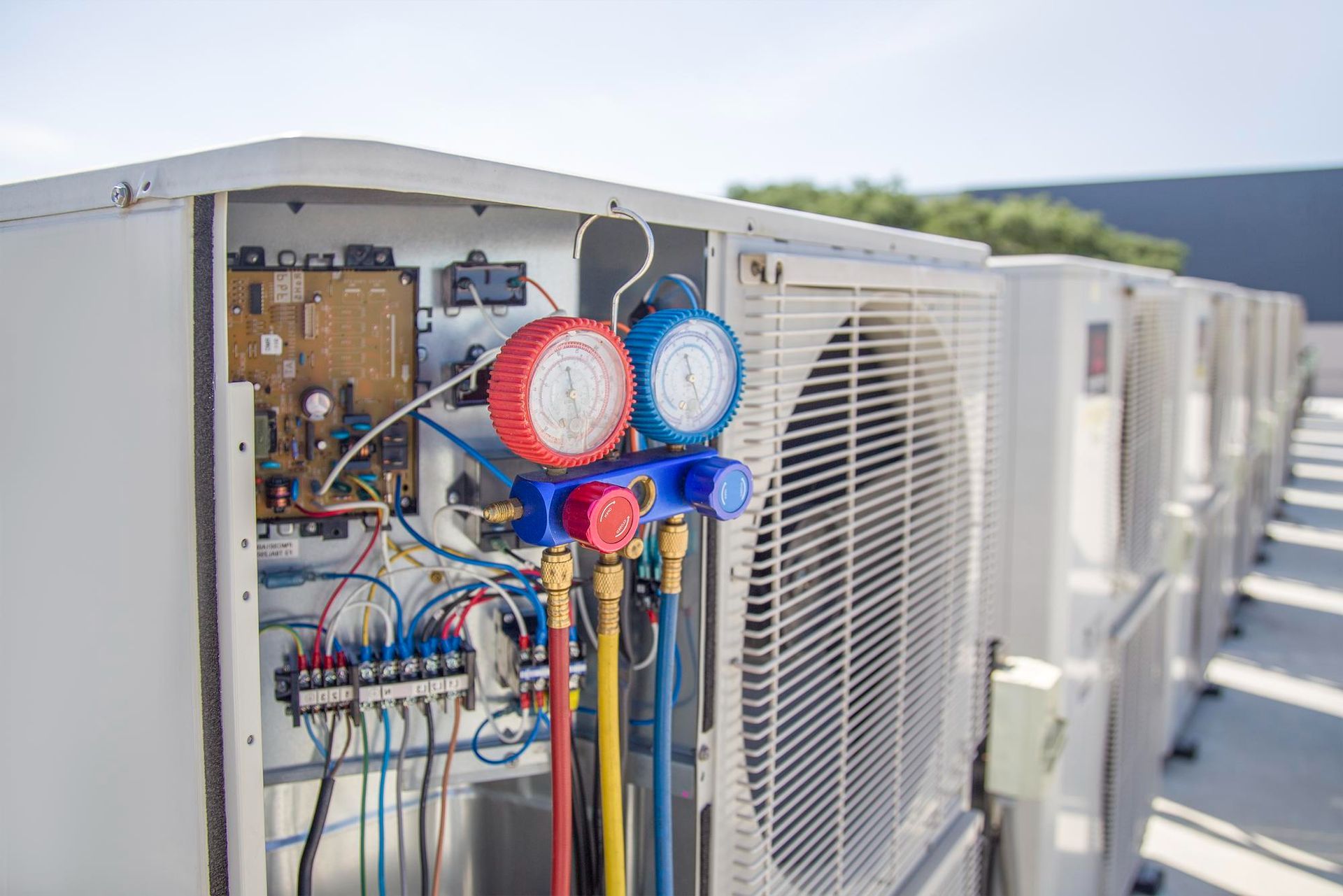 HVAC units with gauges connected, outdoors. One unit's panel is open. Blue and red gauges. White units.