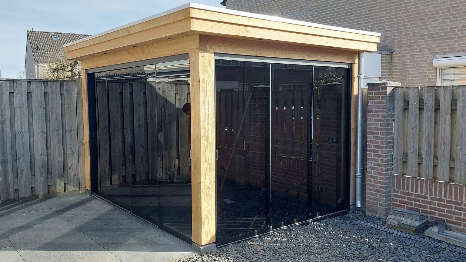 Een houten prieel met glazen schuifdeuren staat voor een bakstenen gebouw.