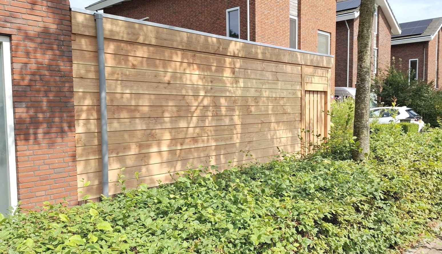 Een hoge, horizontale houten schutting tegen een bakstenen huis met groene struiken op de voorgrond.