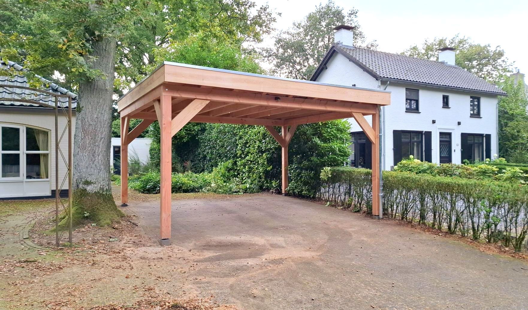 Een houten carport staat op een grindpad naast een wit huis met een donker dak en groene bomen.