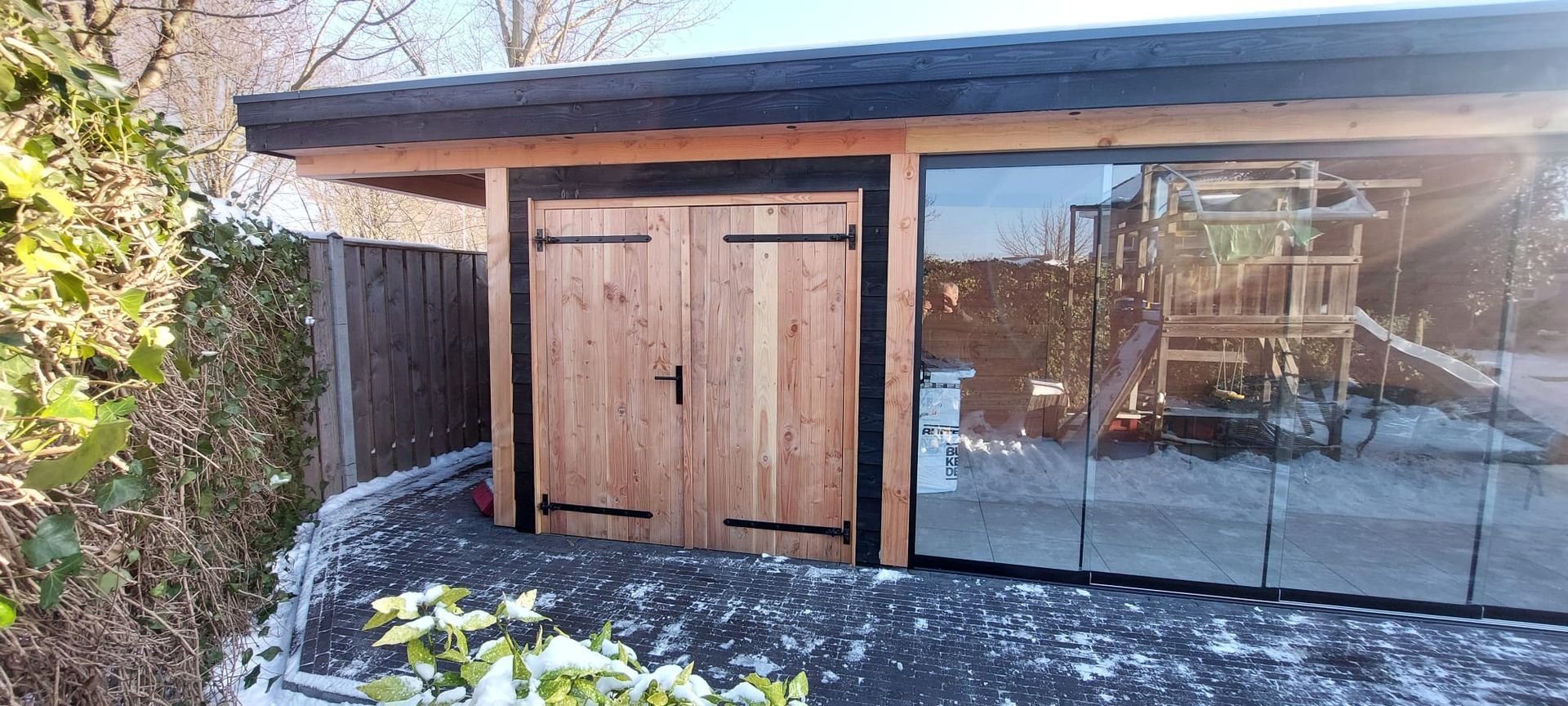 Een moderne tuinconstructie met een dubbele houten deur en grote glazen panelen, geplaatst op een grindoppervlak met een dun laagje sneeuw.