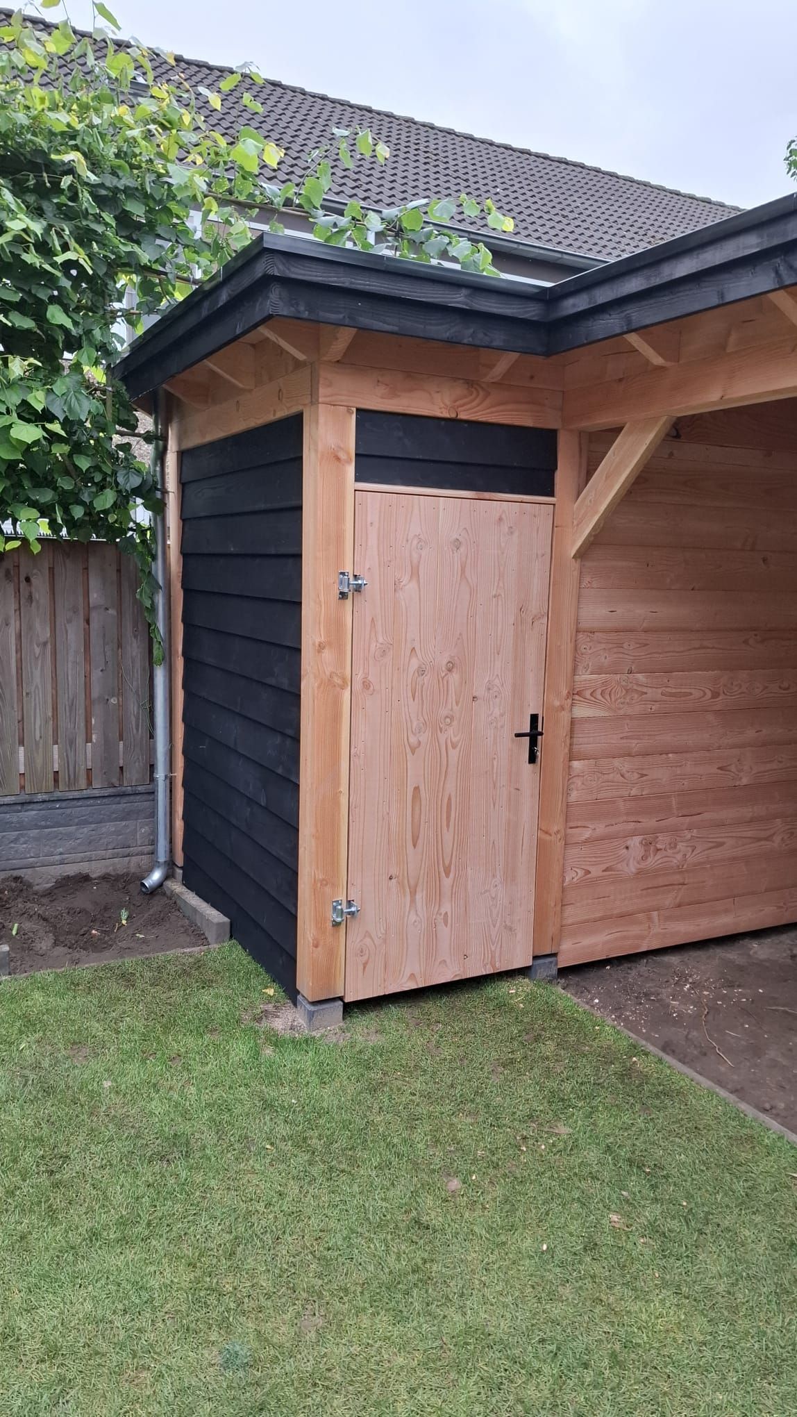 Een onafgewerkte houten deur in de hoek van een tuinhuisje met zwarte horizontale gevelbekleding en een bijpassend dak.