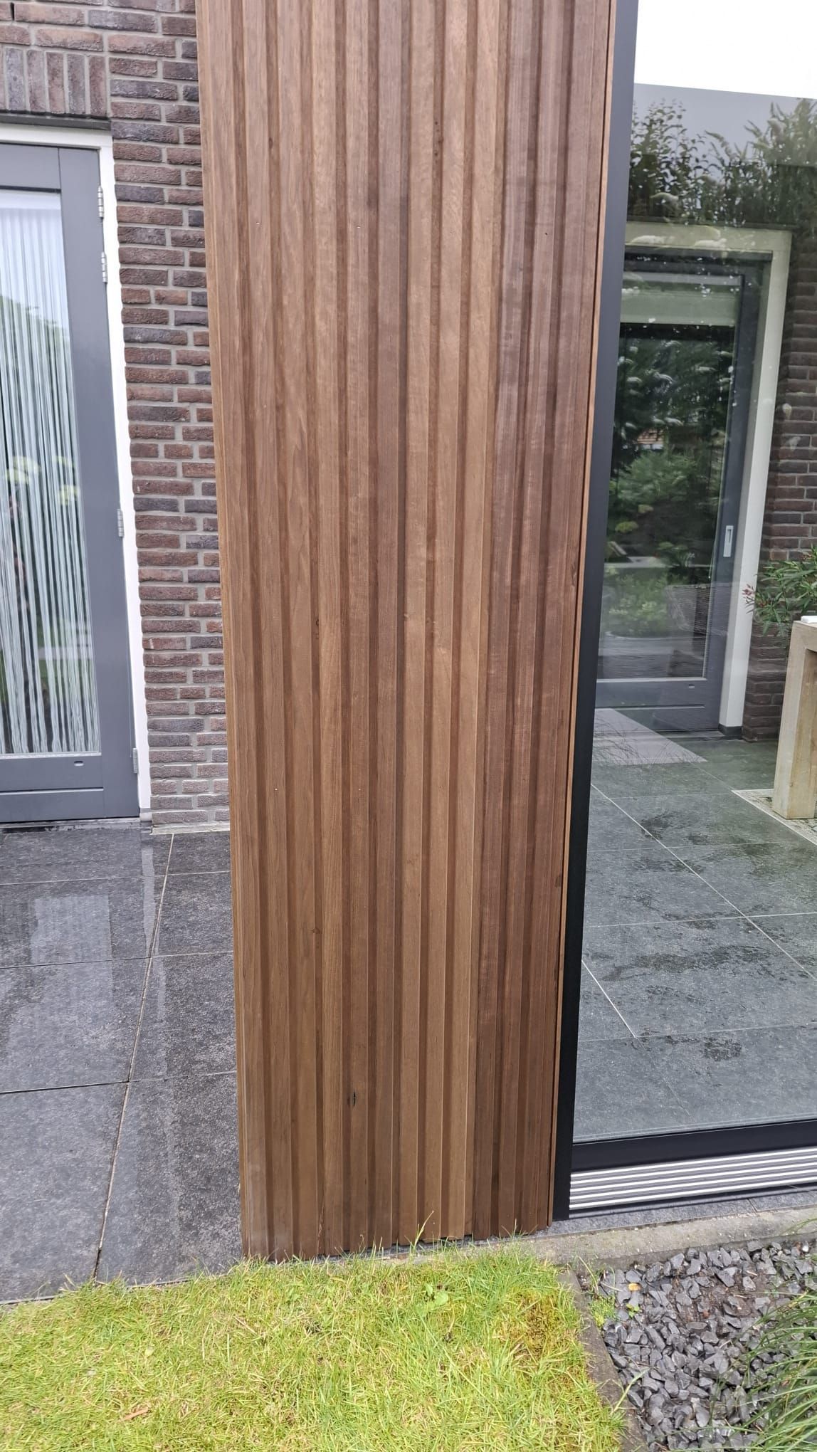 Een verticaal gedeelte van donkerhouten lattenbekleding op de gevel van een modern huis, naast een glazen deur en een stenen terras.