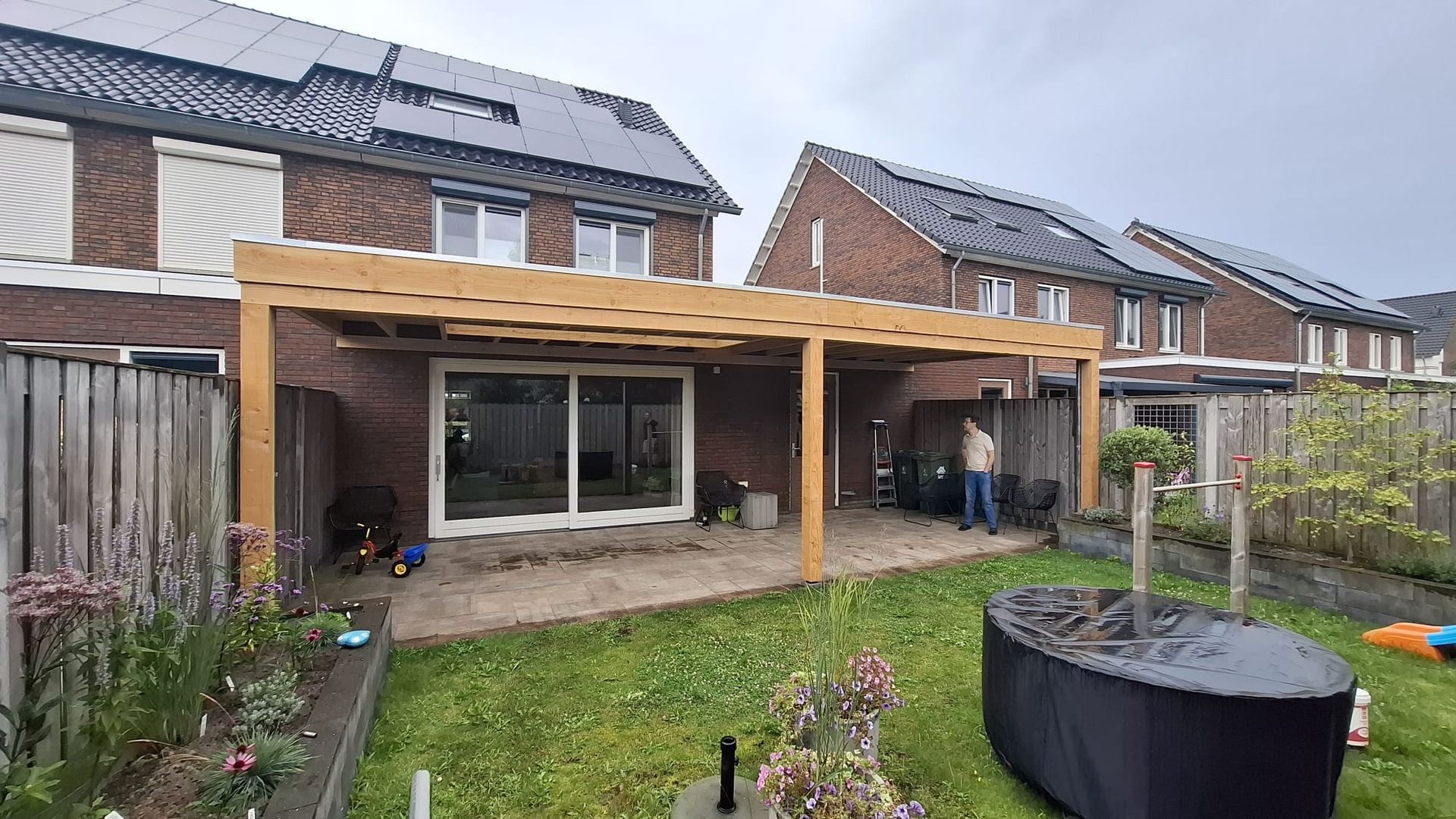 Een houten pergola aan de achterkant van een bakstenen huis, met uitzicht op een grasveld met een overdekte tafel.
