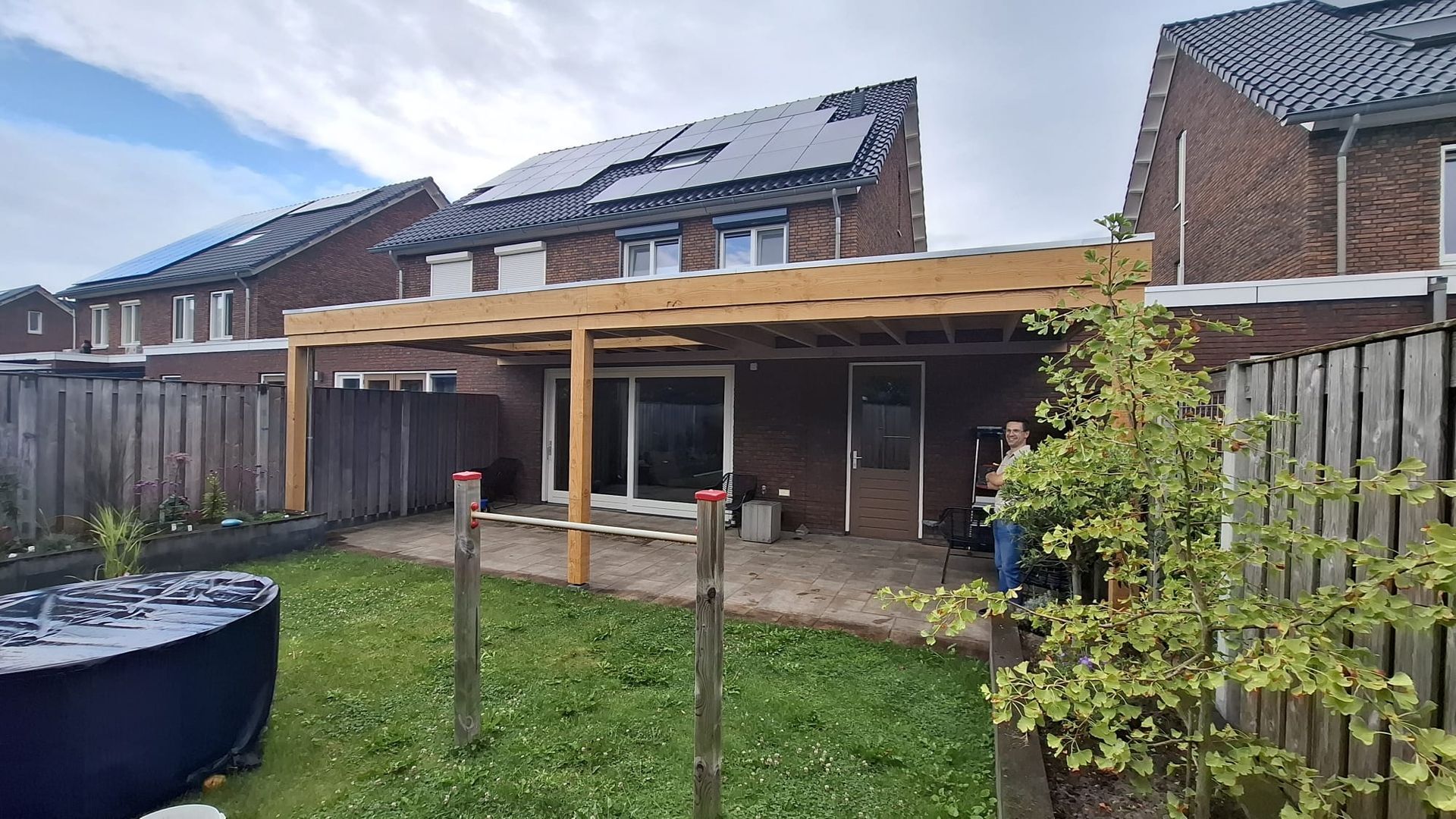 Een houten pergolaframe in aanbouw, bevestigd aan de achtermuur van een huis met zonnepanelen op het dak.