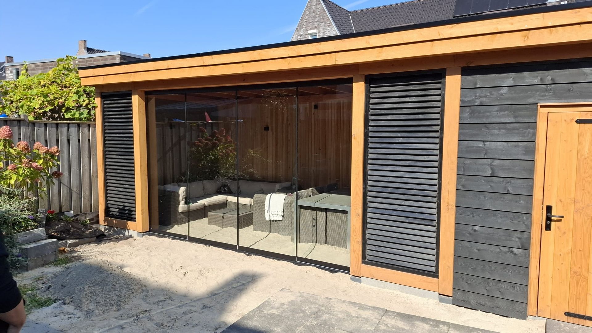 Een houten tuinpaviljoen met glazen schuifdeuren, zwarte latten aan de zijkanten en een gezellige zithoek binnenin.