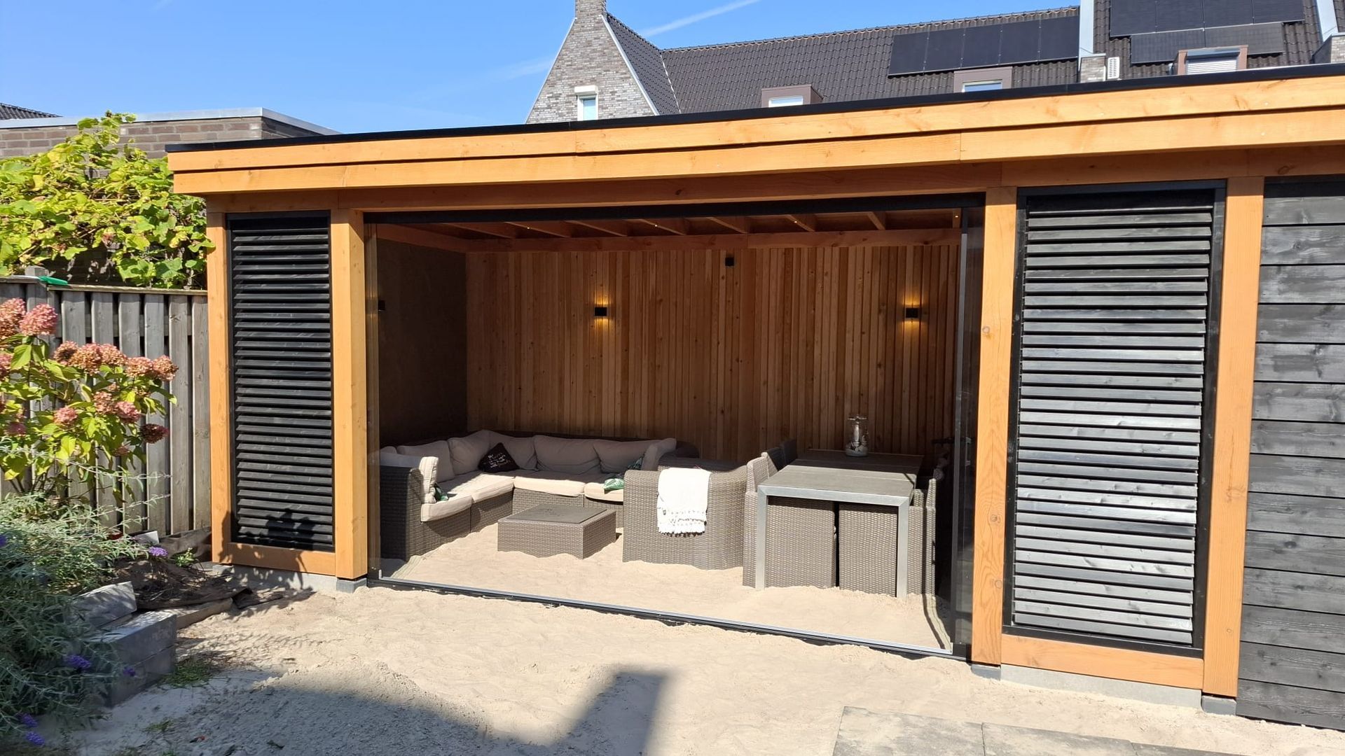 Een modern houten tuinpaviljoen met een loungehoek, rieten meubels, houten lambrisering en decoratieve zwarte schermen.