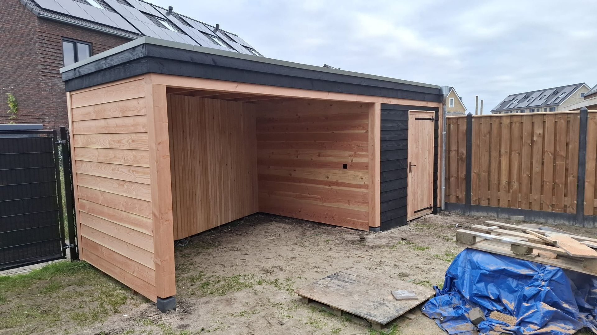 Een gedeeltelijk gebouwd houten tuinhuisje met zwarte afwerking en deur, gelegen in een onafgewerkte woontuin.