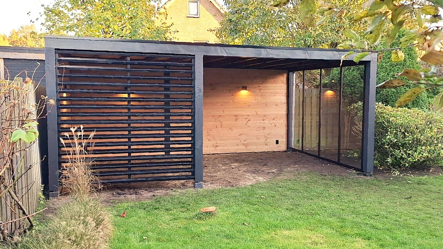 Een moderne tuinconstructie met een wand van zwarte latten, een houten achterwand en een gedeeltelijk met glas afgesloten zijkant.