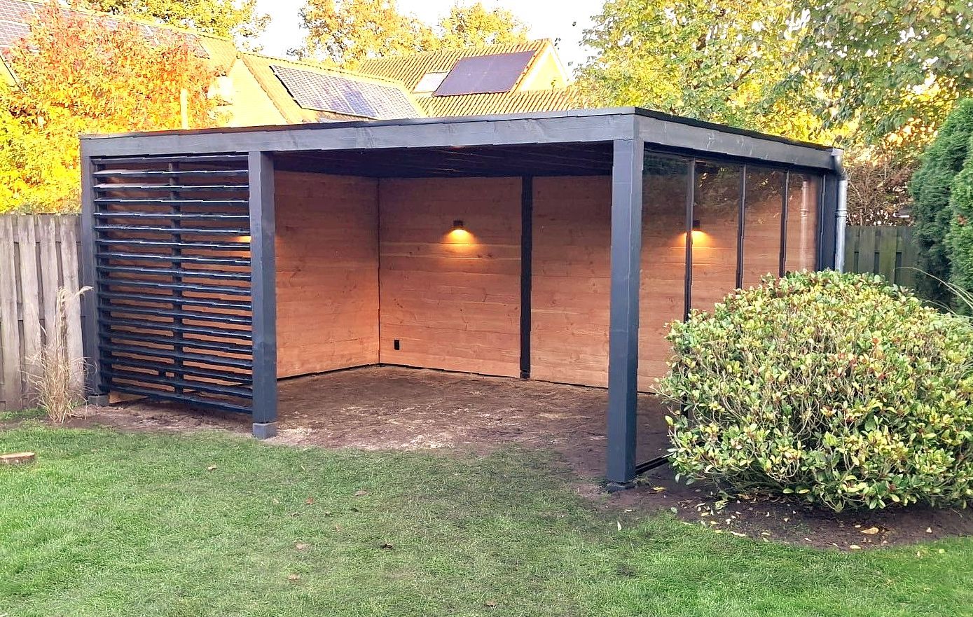Een modern, open tuinpaviljoen met een donker metalen frame, lichte houten lambrisering en verlichte wandlampen op een grasveld.