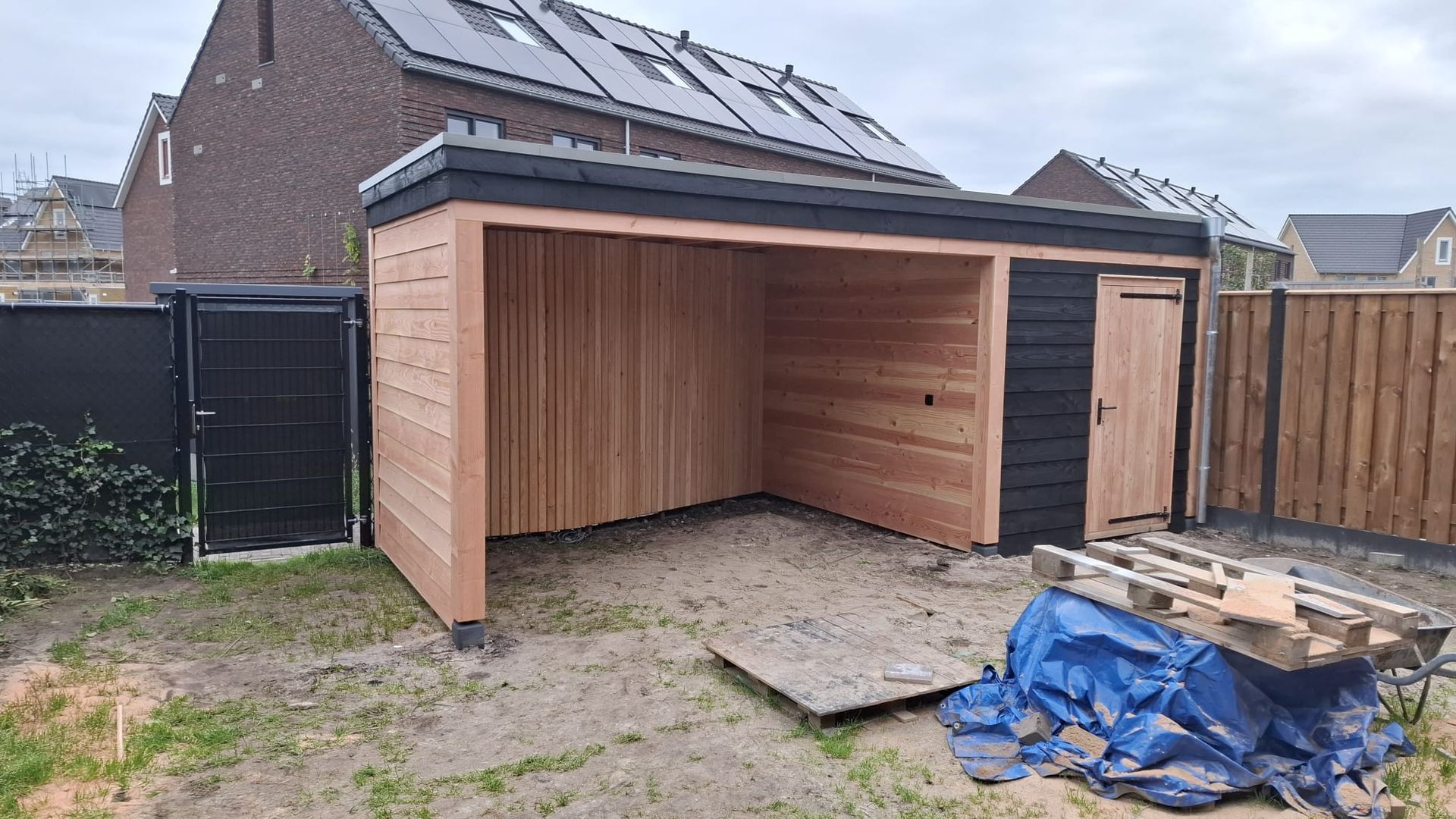 Een houten tuinconstructie met een open overkapping en een gesloten berging, geplaatst in een achtertuin onder een bewolkte hemel.