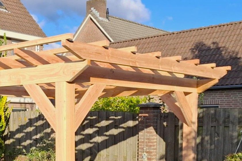 Een houten pergola met spanten en hoekverstevigingen in een achtertuin van een woonhuis.