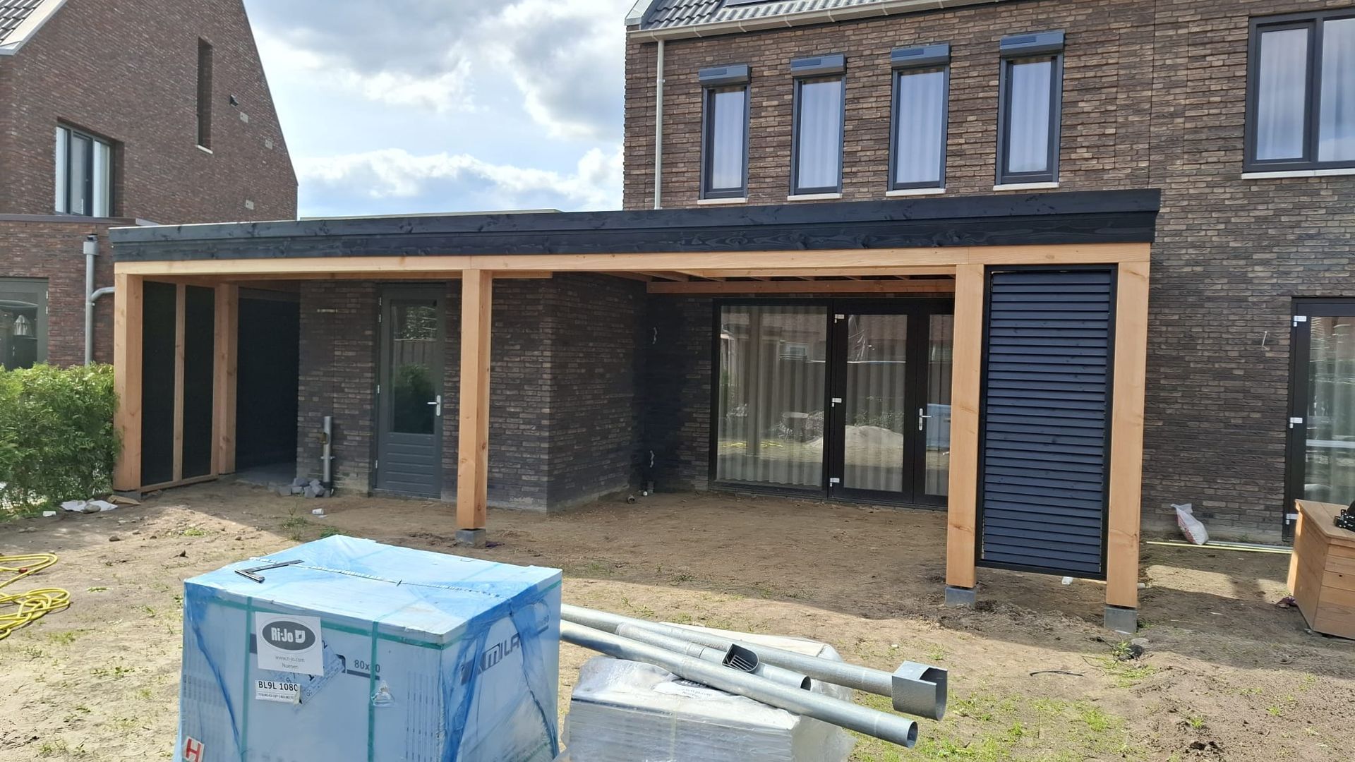 Een houten terrasoverkapping tegen een donker bakstenen huis, met een blauwe opbergbox en metalen buizen op een zanderige binnenplaats.