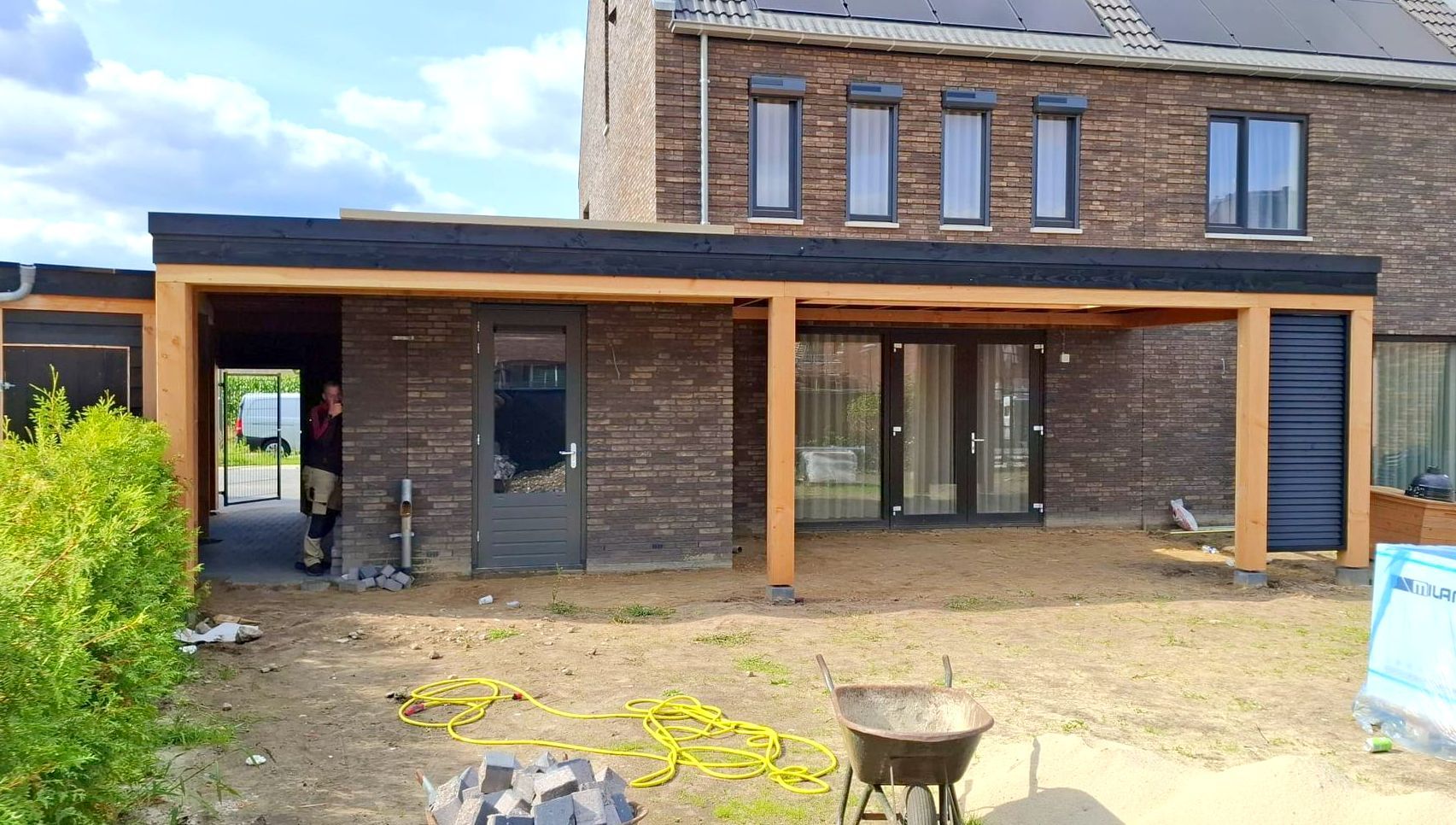 Een achtertuin met een terras, voorzien van een moderne houten pergola die aan een donkerbruin bakstenen huis is bevestigd.