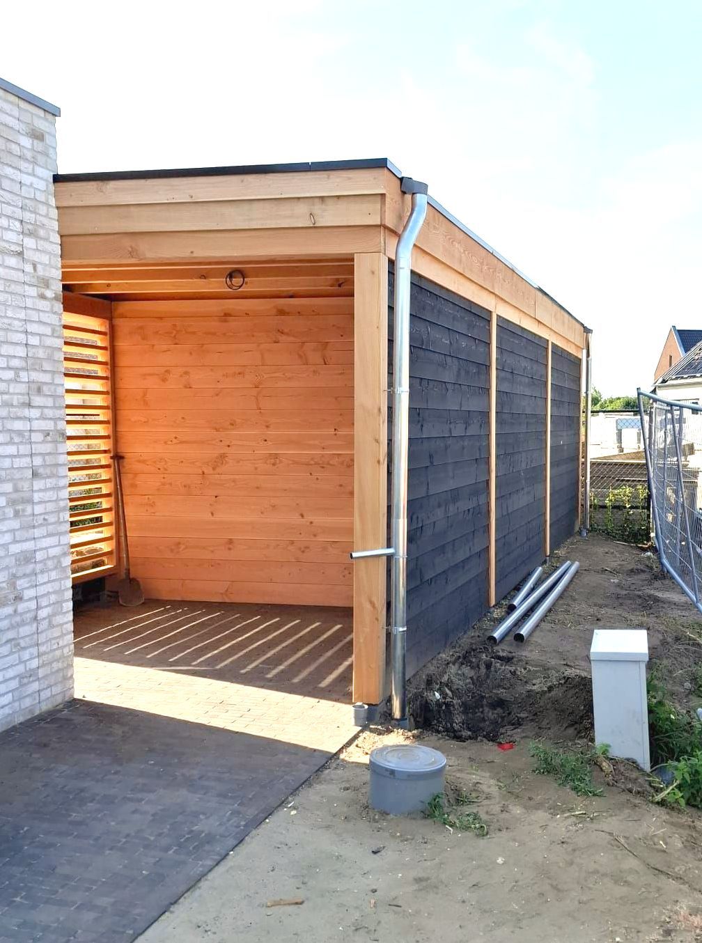 Een moderne carport met een lichte houten binnenkant, donkere houten gevelbekleding en een zilverkleurige regenpijp naast een licht bakstenen huis.