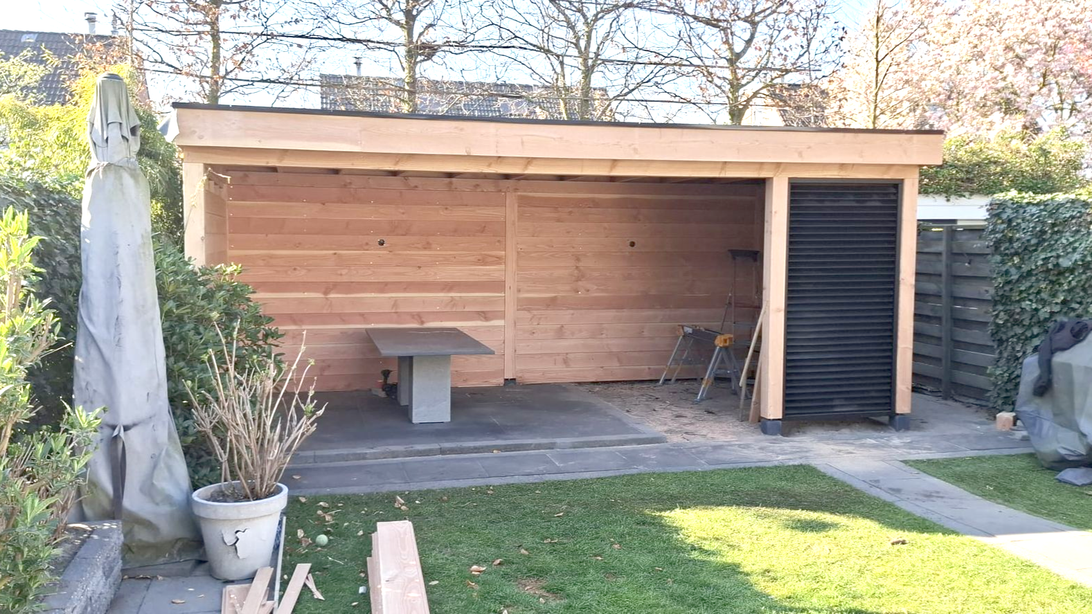 Een houten tuinpaviljoen met een plat dak, een gedeeltelijk overdekt zitgedeelte en een zwart opbergvak aan de rechterkant.