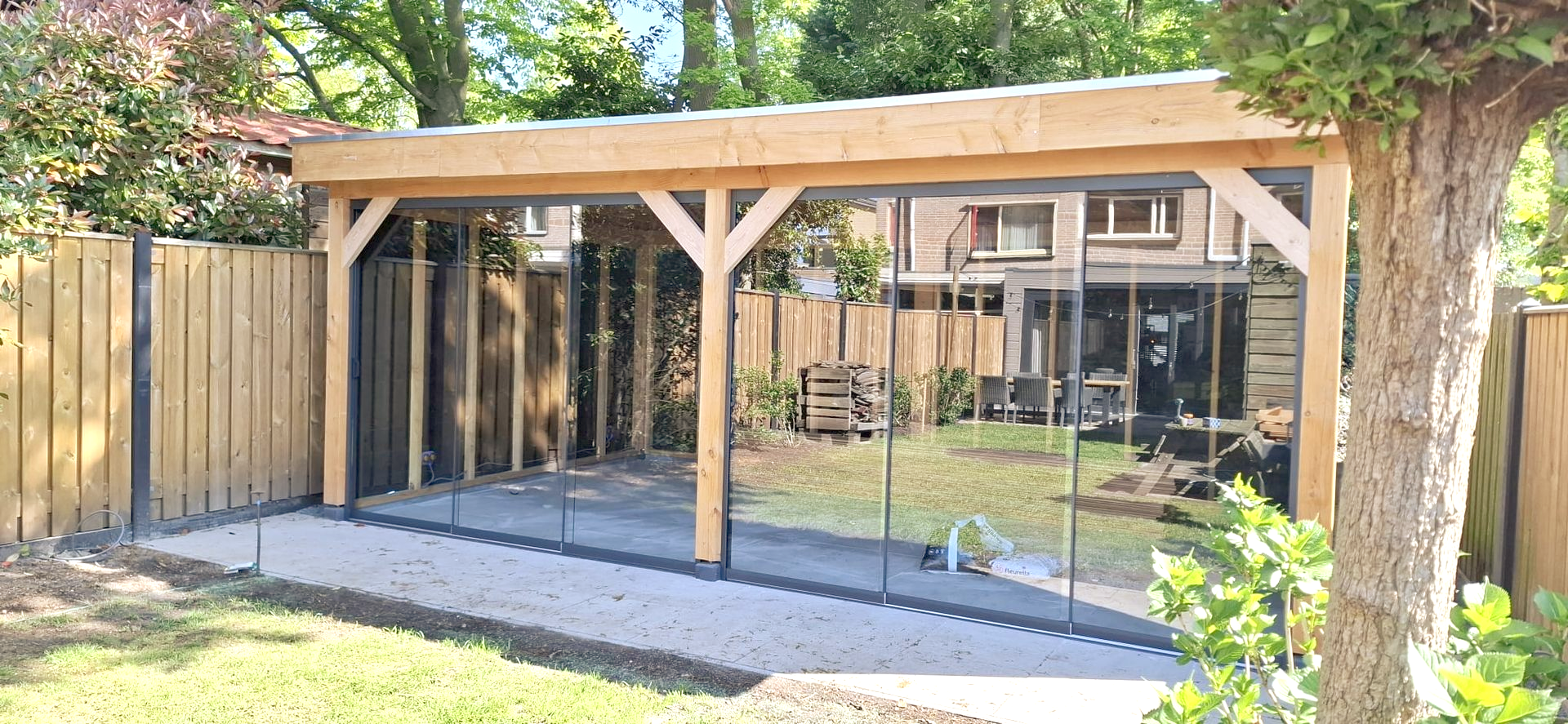Een houten tuinpaviljoen met plat dak en glazen panelen, gelegen aan een grindpad in een omheinde achtertuin.