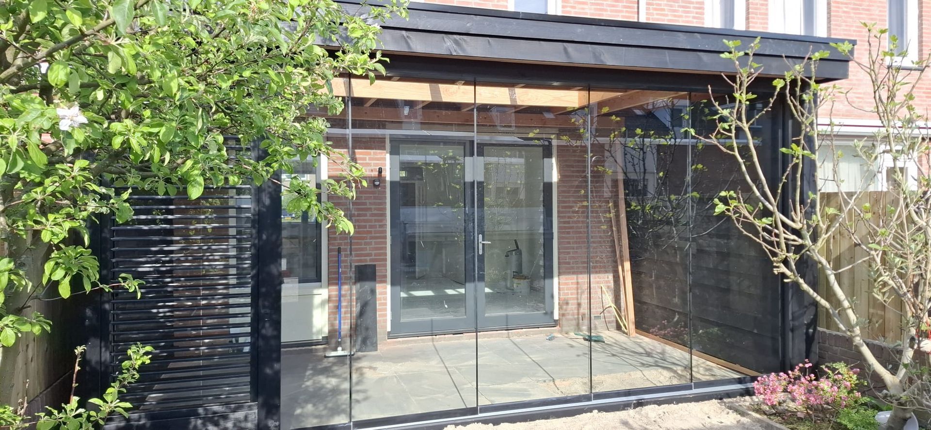 Een moderne tuinkamer met een pannendak, glazen schuifwanden en donkerhouten gevelbekleding, gelegen in een weelderige woontuin.