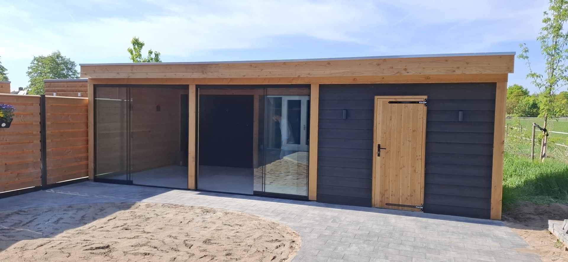 Een moderne tuinkamer met plat dak, schuifdeuren van glas, zwarte horizontale gevelbekleding en een houten deur.