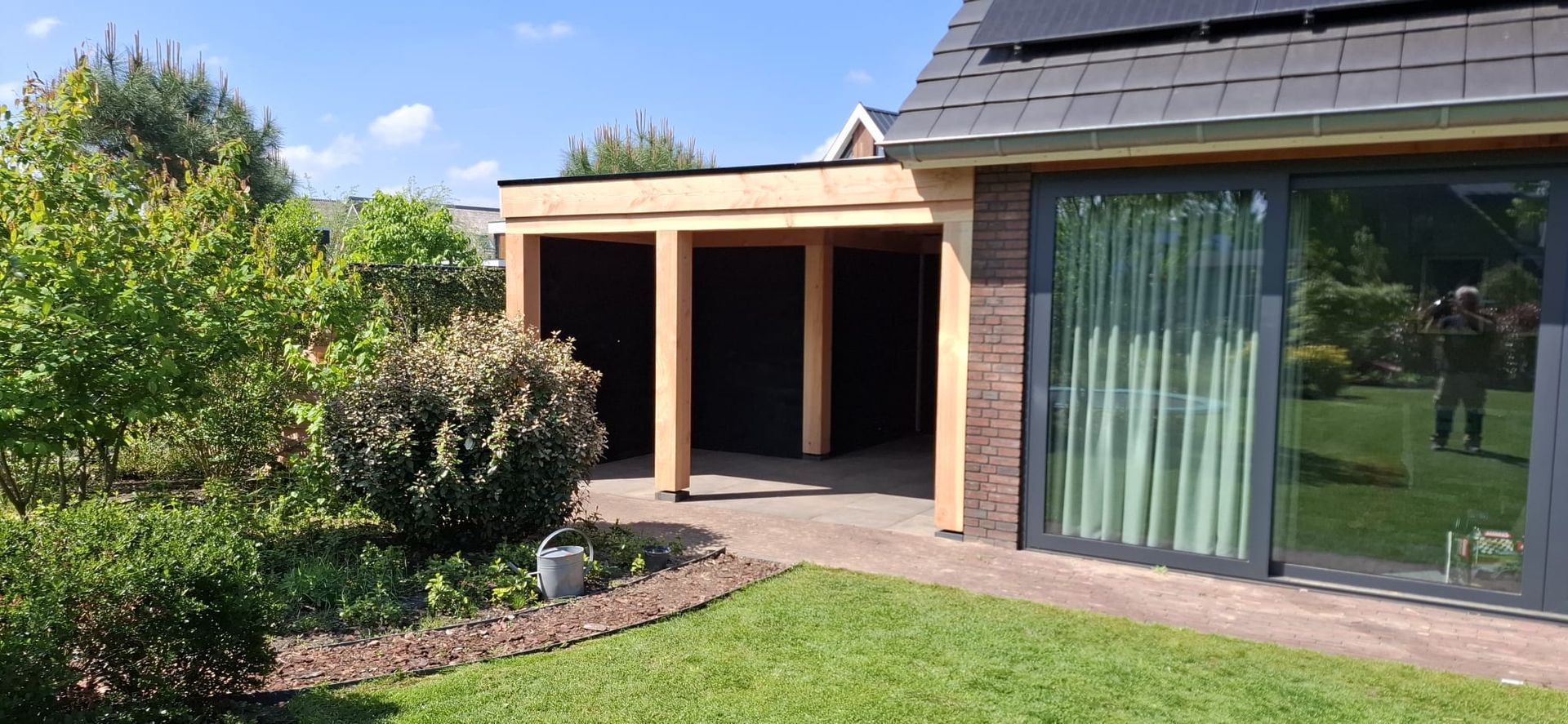 Een houten terrasoverkapping, bevestigd aan een bakstenen huis naast een glazen schuifdeur en een weelderige tuin op een zonnige dag.