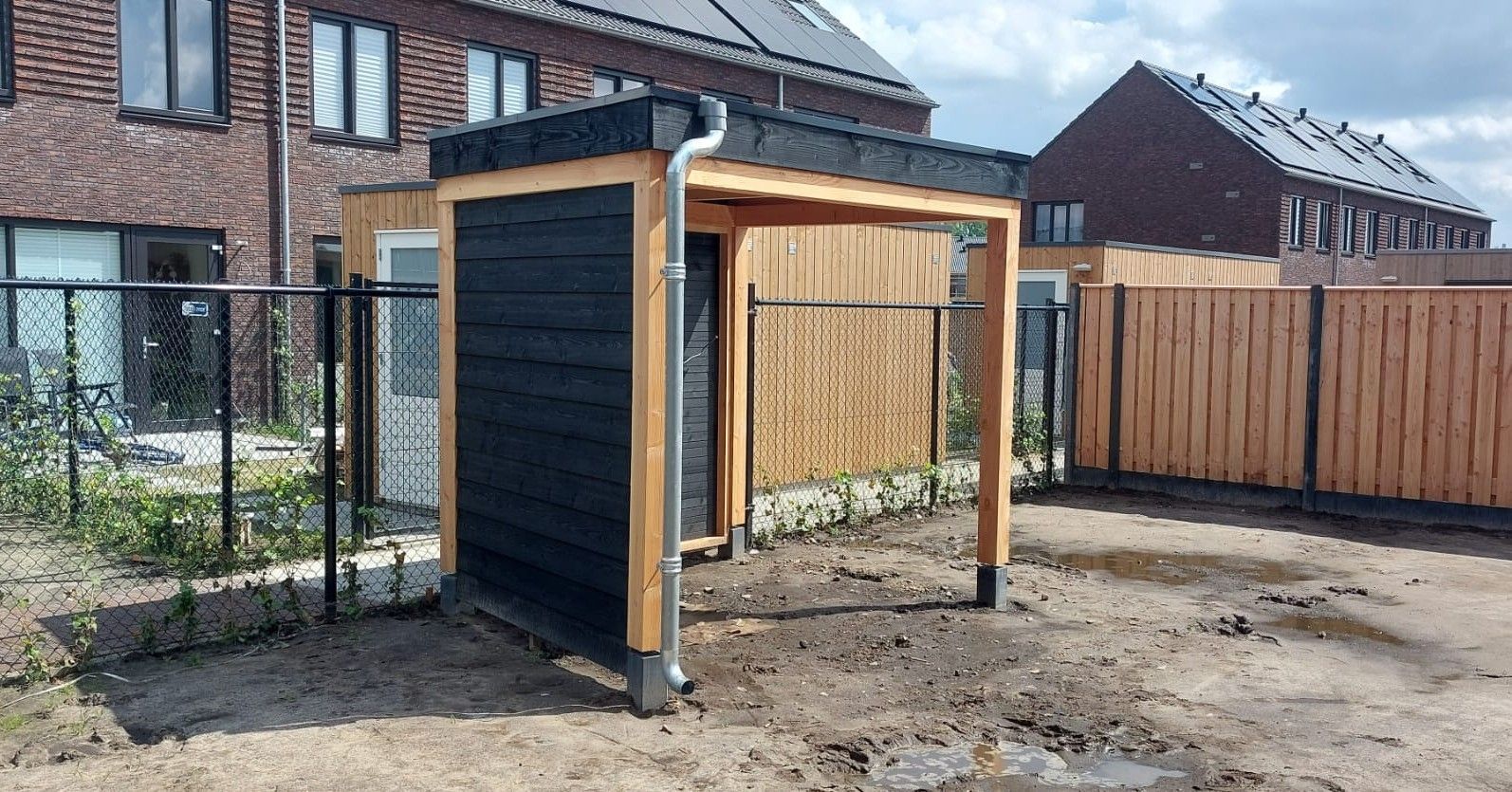 Een klein houten schuurtje staat midden op een zandveld naast een houten hek.