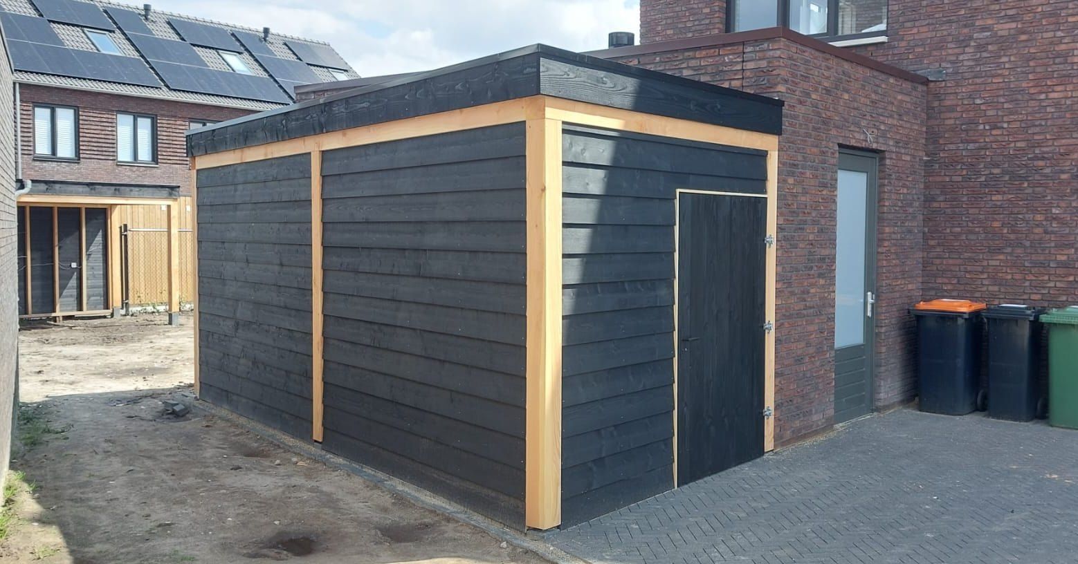 Een zwart houten schuurtje staat naast een bakstenen gebouw.