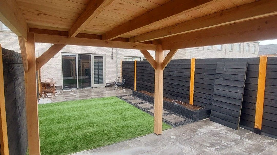 Een houten pergola staat midden in de achtertuin, naast een hek.