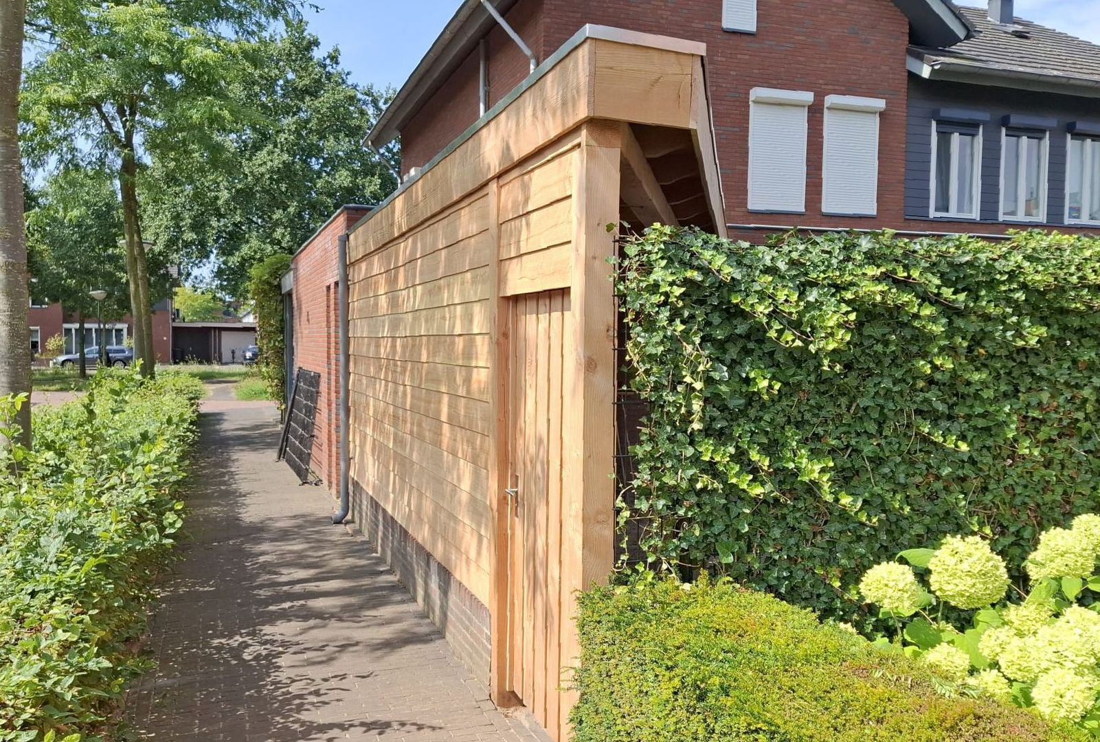 Een nieuwe houten schutting met schuur naast een bakstenen huis, een bestraat pad en een keurig gesnoeide groene haag.