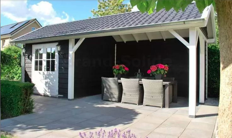 Zwarte tuinschuur met een overdekt terras, voorzien van een eethoek met rieten stoelen en rode bloemen op een stenen terras.