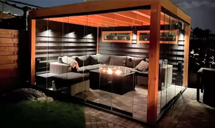 Een verlicht houten tuinpaviljoen met glazen wanden, met loungebanken en een vuurplaats op een stenen terras 's avonds.